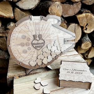 May include: Wooden wedding guest book shaped like the Millennium Falcon from Star Wars. The book has a slot for guests to drop in wooden hearts. The box is engraved with the names "Eva & Alexander" and the date "14.10.2024".