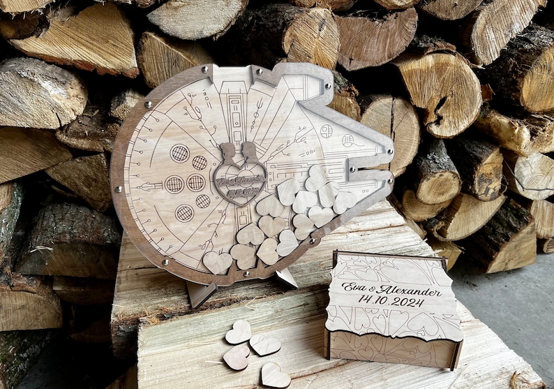 May include: Wooden wedding guest book shaped like the Millennium Falcon from Star Wars. The book has a slot for guests to drop in wooden hearts. The box is engraved with the names "Eva & Alexander" and the date "14.10.2024".