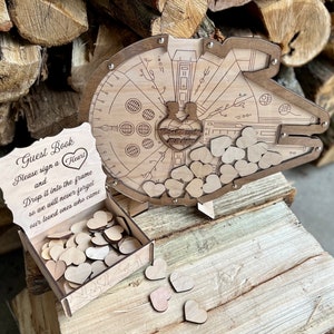 May include: Wooden guest book shaped like the Millennium Falcon from Star Wars. The guest book has a slot for guests to drop wooden hearts into. The sign reads "Guest Book Please sign a Heart and Drop it into the frame so we will never forget our loved ones who came."