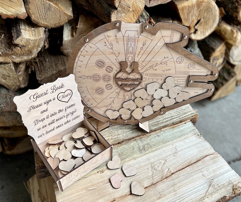 May include: Wooden guest book shaped like the Millennium Falcon from Star Wars. The guest book has a slot for guests to drop wooden hearts into. The sign reads "Guest Book Please sign a Heart and Drop it into the frame so we will never forget our loved ones who came."