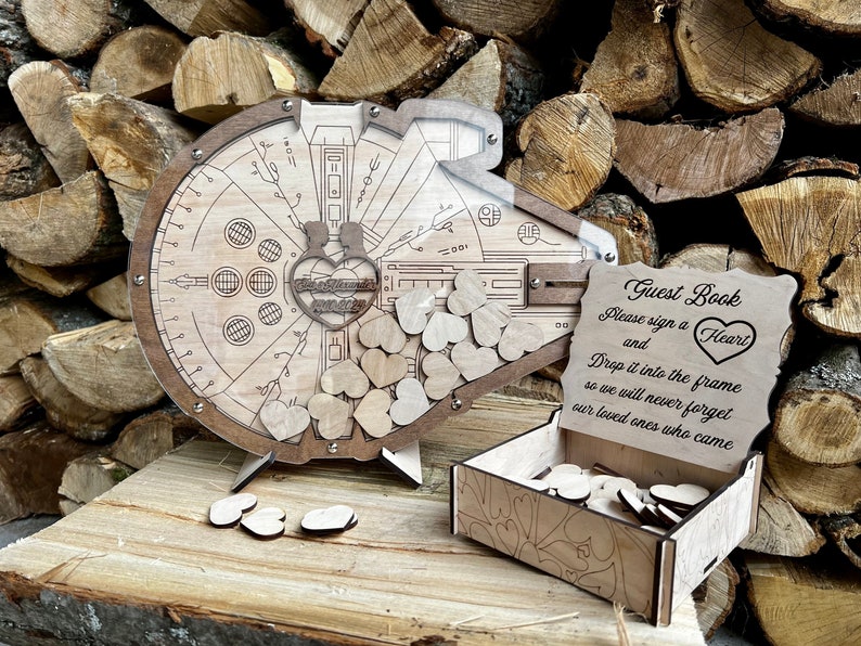 May include: Wooden guest book shaped like a spaceship with a heart-shaped opening. The sign reads "Guest Book Please sign a Heart and Drop it into the frame so we will never forget our loved ones who came."
