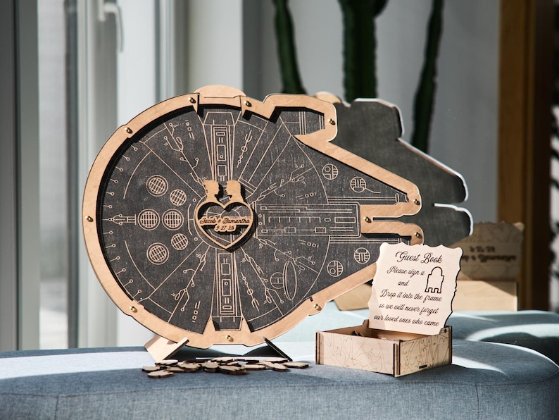 May include: A wooden guest book shaped like the Millennium Falcon from Star Wars. The guest book has a heart-shaped opening in the center where guests can drop their well wishes. The sign reads "Guest Book Please sign a and Drop it into the frame so we will never forget our loved ones who came"