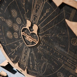May include: A wooden cutout of a couple in a heart shape with the text "Jacob & Samantha 9.27.23" on a black background with a detailed design of a spaceship.