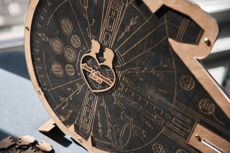 May include: A wooden cutout of a couple in a heart shape with the text "Jacob & Samantha 9.27.23" on a black background with a detailed design of a spaceship.