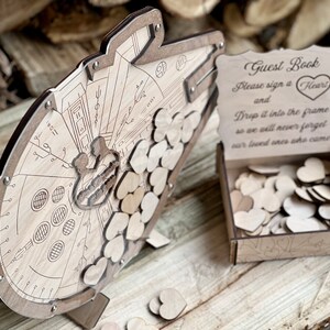 May include: A wooden guest book shaped like the Millennium Falcon from Star Wars. The guest book has a slot for guests to drop wooden hearts into. The sign reads "Guest Book, Please sign a Heart and Drop it into the frame so we will never forget our loved ones who came."