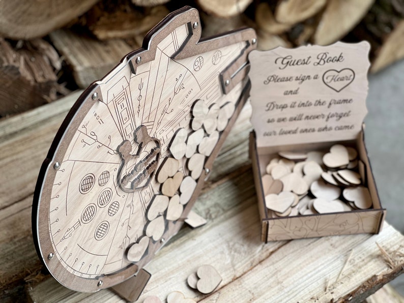May include: A wooden guest book shaped like the Millennium Falcon from Star Wars. The guest book has a slot for guests to drop wooden hearts into. The sign reads "Guest Book, Please sign a Heart and Drop it into the frame so we will never forget our loved ones who came."