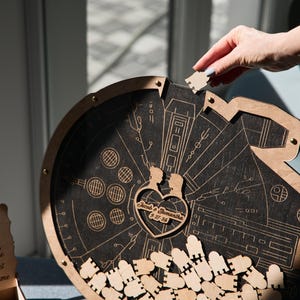May include: A wooden guest book shaped like the Millennium Falcon from Star Wars. The guest book has a heart-shaped cutout with the names "Jared & Samantha" and the date "05.27.2023" engraved on it. The guest book is filled with small wooden pieces that guests can sign and drop into the cutout.