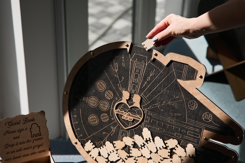 May include: A wooden guest book shaped like the Millennium Falcon from Star Wars. The guest book has a heart-shaped cutout with the names "Jared & Samantha" and the date "05.27.2023" engraved on it. The guest book is filled with small wooden pieces that guests can sign and drop into the cutout.