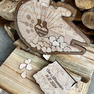 May include: Wooden wedding card box shaped like the Millennium Falcon from Star Wars. The box is decorated with hearts and the text "Eva & Alexander 14.10.2024".
