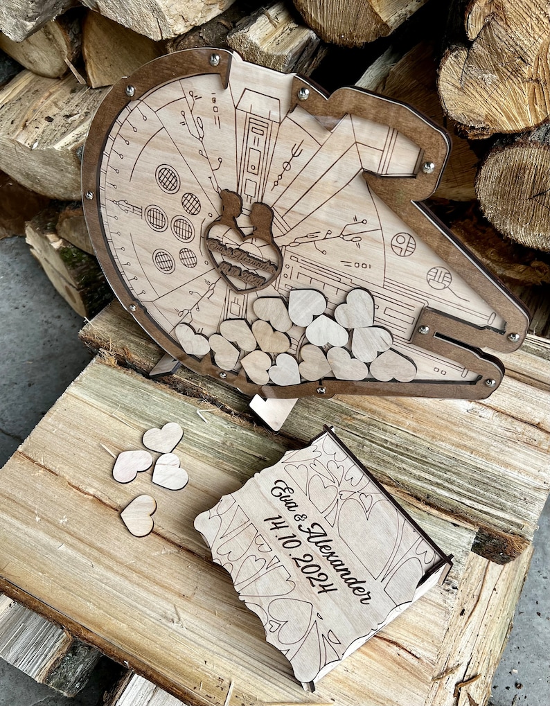 May include: Wooden wedding card box shaped like the Millennium Falcon from Star Wars. The box is decorated with hearts and the text "Eva & Alexander 14.10.2024".