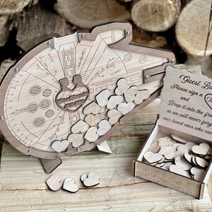 May include: A wooden guest book shaped like the Millennium Falcon from Star Wars. The guest book has a slot for guests to drop wooden hearts into. The sign reads "Guest Book Please sign a Heart and Drop it into the frame so we will never forget our loved ones who came."