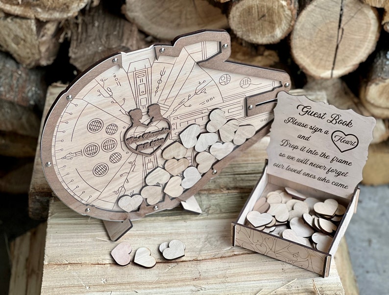 May include: A wooden guest book shaped like the Millennium Falcon from Star Wars. The guest book has a slot for guests to drop wooden hearts into. The sign reads "Guest Book Please sign a Heart and Drop it into the frame so we will never forget our loved ones who came."