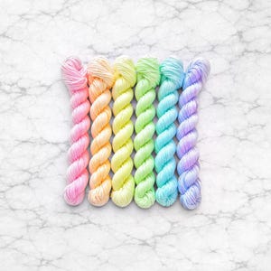 Könnte beinhalten: Sechs Stränge farbigen Garns in einer Reihe. Die Garnfarben sind Rosa, Pfirsich, Gelb, Grün, Blau und Lila. Das Garn wird auf einer weißen Marmoroberfläche präsentiert.