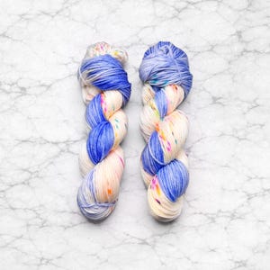 May include: Two skeins of yarn with a cream base, vibrant blue sections, and colorful speckles. The yarn is twisted and arranged in a vertical position against a marble background. The blue sections are a darker shade at the top and bottom.