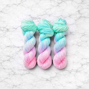 Pode incluir: Tr&ecirc;s novelos de fio multicolorido. O fio apresenta um efeito degrad&ecirc;, passando do rosa ao azul e depois ao verde menta. O fio &eacute; apresentado sobre uma superf&iacute;cie de m&aacute;rmore branco. Cada novelo pesa aproximadamente 100 gramas.
