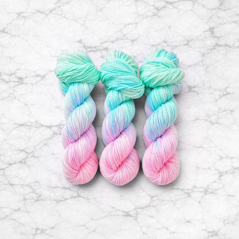 Pode incluir: Tr&ecirc;s novelos de fio multicolorido. O fio apresenta um efeito degrad&ecirc;, passando do rosa ao azul e depois ao verde menta. O fio &eacute; apresentado sobre uma superf&iacute;cie de m&aacute;rmore branco. Cada novelo pesa aproximadamente 100 gramas.