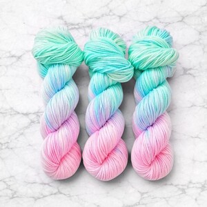 Pode incluir: Tr&ecirc;s novelos de fio tingido &agrave; m&atilde;o em um gradiente de cores. O fio transita de um verde menta vibrante no topo, por tons de azul, para um rosa suave na parte inferior. O fio &eacute; torcido e disposto sobre uma superf&iacute;cie de m&aacute;rmore branco.