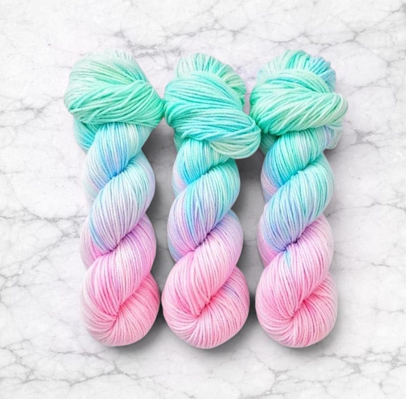 Pode incluir: Tr&ecirc;s novelos de fio tingido &agrave; m&atilde;o em um gradiente de cores. O fio transita de um verde menta vibrante no topo, por tons de azul, para um rosa suave na parte inferior. O fio &eacute; torcido e disposto sobre uma superf&iacute;cie de m&aacute;rmore branco.