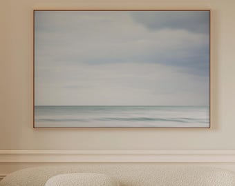 May include: A cream-coloured, rounded, textured sofa with a matching bolster cushion, a small black three-legged stool, and a large rectangular framed landscape print depicting a calm sea under a cloudy sky, all set in a neutral-toned room.