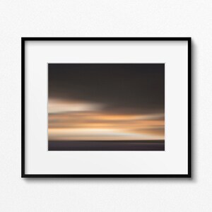 May include: A framed photograph of a blurred sunset over a body of water. The sky is a gradient of orange, yellow, and purple, with a few wispy clouds. The water is a dark blue, and the horizon is a thin line.