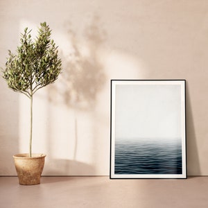 May include: A framed art print with a black border depicts a serene ocean scene with a misty horizon. Beside it, a small potted olive tree stands, its green leaves contrasting with the neutral tones of the wall and floor. The room is bathed in soft, natural light.