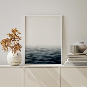 May include: A framed print of the ocean. The image shows a tranquil view of the sea, with dark blue water and a hazy sky. The print is displayed on a white cabinet, with decorative items like a vase and books.