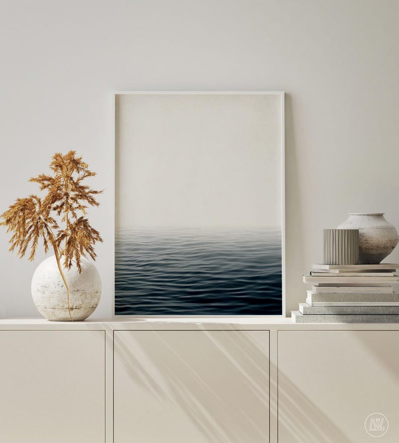 May include: A framed print of the ocean. The image shows a tranquil view of the sea, with dark blue water and a hazy sky. The print is displayed on a white cabinet, with decorative items like a vase and books.
