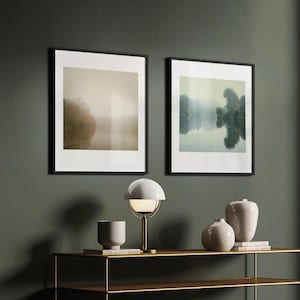 May include: Two framed landscape photographs of a misty lake with trees in the background. A white lamp with a gold base sits on a gold metal shelf with two white ceramic vases and a book.