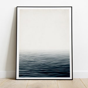 May include: A framed art print featuring a minimalist seascape. The image shows a gradient of colors, transitioning from a bright white sky to a deep blue ocean. The water's surface is gently rippled, creating a sense of calm and serenity.
