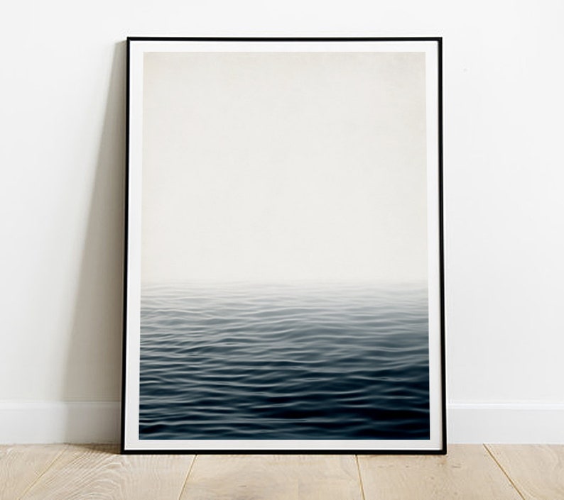 May include: A framed art print featuring a minimalist seascape. The image shows a gradient of colors, transitioning from a bright white sky to a deep blue ocean. The water's surface is gently rippled, creating a sense of calm and serenity.