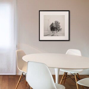 May include: A black and white photograph of a snowy landscape with a windmill and trees in the distance. The photo is framed in a black frame and is hanging on a wall above a white table with four white chairs.
