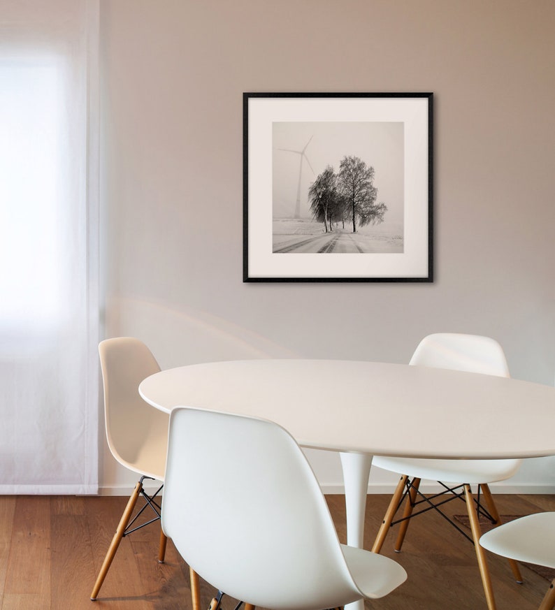 May include: A black and white photograph of a snowy landscape with a windmill and trees in the distance. The photo is framed in a black frame and is hanging on a wall above a white table with four white chairs.