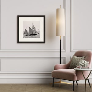 May include: A black and white photograph of sailboats on the water, framed in a black frame and hanging on a white wall. A pink armchair with a throw pillow sits in front of the framed photo.
