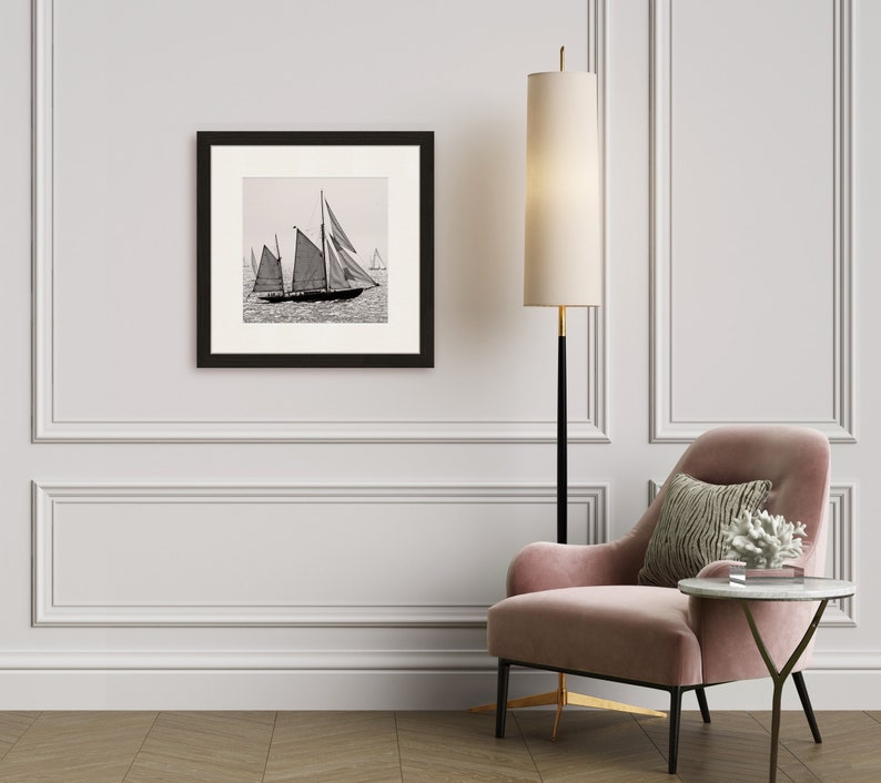 May include: A black and white photograph of sailboats on the water, framed in a black frame and hanging on a white wall. A pink armchair with a throw pillow sits in front of the framed photo.
