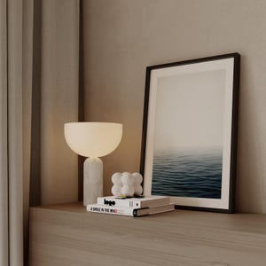 May include: A modern interior scene featuring a white table lamp with a marble base, a framed picture of the ocean, and two books. The books have the text "logo" and "A SMILE IN THE MIND". A decorative white sculpture is also present.