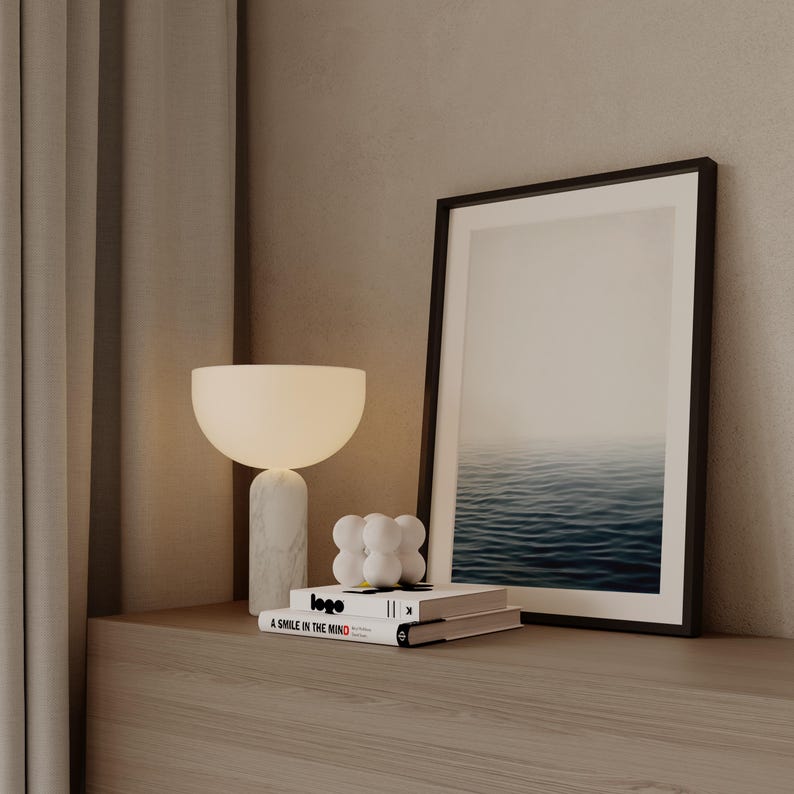 May include: A modern interior scene featuring a white table lamp with a marble base, a framed picture of the ocean, and two books. The books have the text "logo" and "A SMILE IN THE MIND". A decorative white sculpture is also present.