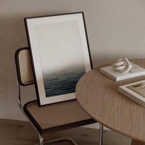 May include: A framed art print depicting a seascape with a gradient sky and rippling water. The artwork is propped against a chair with a woven back and a wooden table with books and a decorative knot. The color palette is neutral.