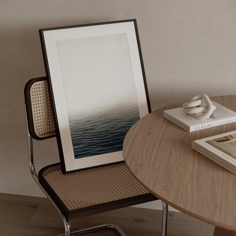 May include: A framed art print depicting a seascape with a gradient sky and rippling water. The artwork is propped against a chair with a woven back and a wooden table with books and a decorative knot. The color palette is neutral.