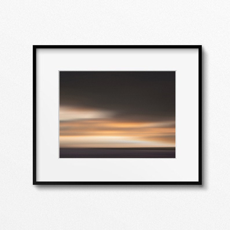 May include: A framed photograph of a blurred sunset over a body of water. The sky is a gradient of orange, yellow, and purple, with a few wispy clouds. The water is a dark blue, and the horizon is a thin line.