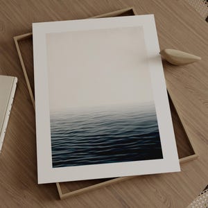 May include: A framed art print featuring a serene ocean scene. The artwork depicts a gradient of colors, transitioning from a misty, light beige sky to deep blue water. The print is set in a light wood frame, resting on a wooden surface.