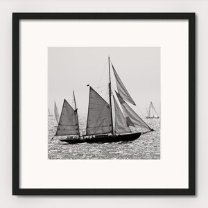 May include: A black and white photograph of a sailboat with its sails billowing in the wind. The sailboat is in the foreground, with other sailboats in the background. The water is choppy and the sky is cloudy.