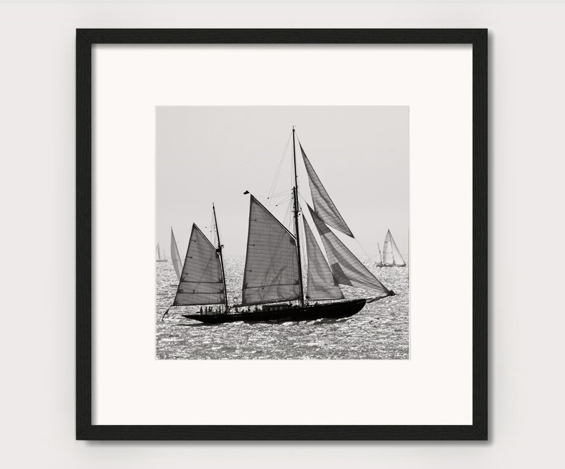 May include: A black and white photograph of a sailboat with its sails billowing in the wind. The sailboat is in the foreground, with other sailboats in the background. The water is choppy and the sky is cloudy.