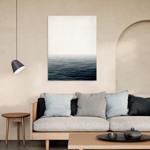 May include: A minimalist living room featuring a canvas print of ocean waves. The room includes a light-colored sofa with throw pillows, a wooden coffee table, and a side table. A black pendant lamp hangs from the ceiling, and the walls are painted in a neutral tone.