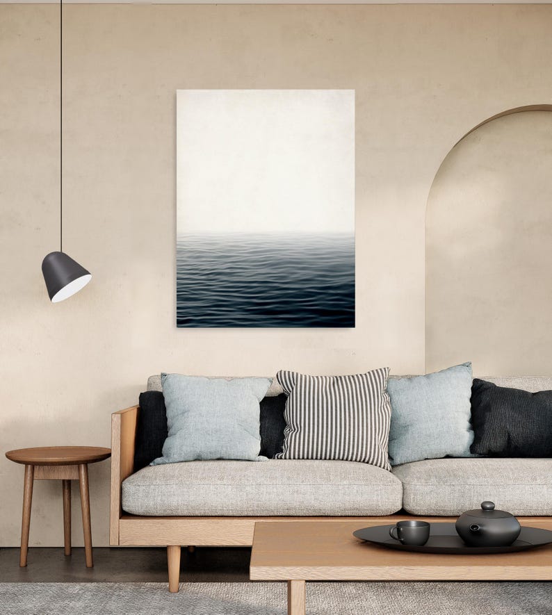 May include: A minimalist living room featuring a canvas print of ocean waves. The room includes a light-colored sofa with throw pillows, a wooden coffee table, and a side table. A black pendant lamp hangs from the ceiling, and the walls are painted in a neutral tone.