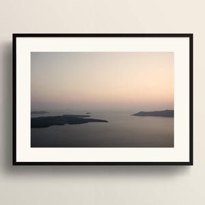 May include: A framed landscape photograph featuring a calm body of water surrounded by dark, silhouetted landmasses under a soft, hazy sky with a gradient of pale peach and light grey tones. The artwork is enclosed in a thin, black rectangular frame.