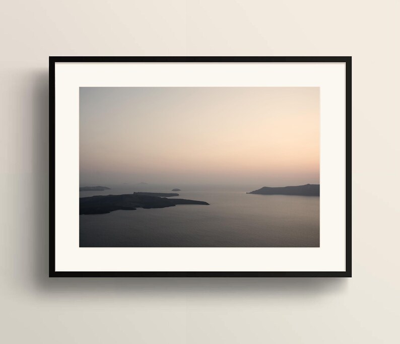 May include: A framed landscape photograph featuring a calm body of water surrounded by dark, silhouetted landmasses under a soft, hazy sky with a gradient of pale peach and light grey tones. The artwork is enclosed in a thin, black rectangular frame.