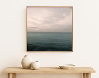 May include: A square framed wall art print depicting a calm ocean horizon under a soft, overcast sky with muted blue and grey tones. The artwork is displayed in a thin wooden frame, capturing a serene seascape with a smooth water surface.
