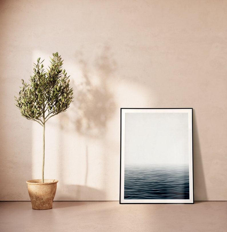 May include: A framed art print with a black border depicts a serene ocean scene with a misty horizon. Beside it, a small potted olive tree stands, its green leaves contrasting with the neutral tones of the wall and floor. The room is bathed in soft, natural light.