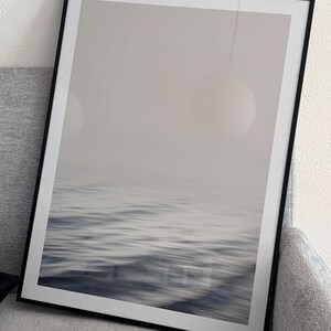 May include: A framed print featuring a blurred seascape in shades of gray and blue. The artwork captures the movement of water with a soft, abstract quality. The black frame adds a modern touch to the minimalist ocean-themed art.