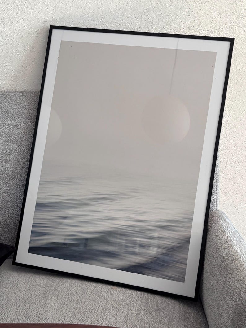 May include: A framed print featuring a blurred seascape in shades of gray and blue. The artwork captures the movement of water with a soft, abstract quality. The black frame adds a modern touch to the minimalist ocean-themed art.
